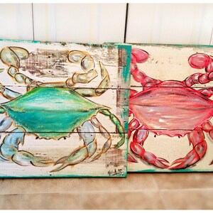 May include: Two painted wooden signs featuring crabs. The left sign has a blue crab with green accents on a whitewashed wood background. The right sign has a red crab on a whitewashed wood background. Both signs have a turquoise border.