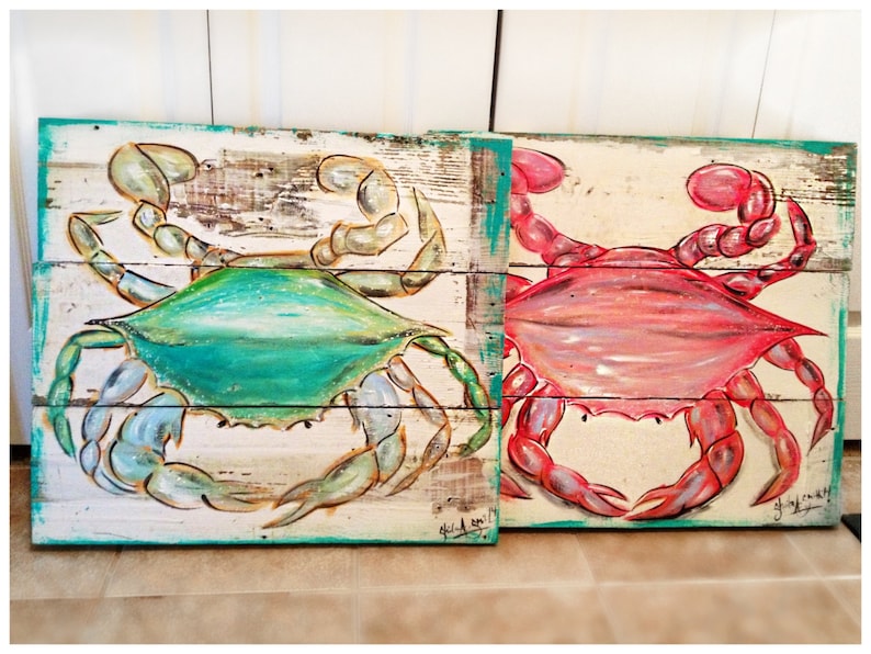 May include: Two painted wooden signs featuring crabs. The left sign has a blue crab with green accents on a whitewashed wood background. The right sign has a red crab on a whitewashed wood background. Both signs have a turquoise border.