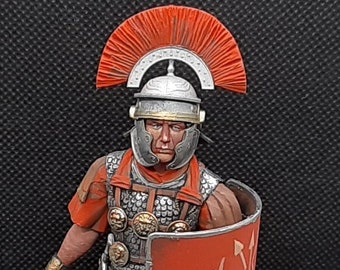 May include: A detailed miniature figurine of a Roman soldier wearing a red tunic, helmet, and armor, holding a sword and a red shield with white markings. The figurine is standing on a black base.