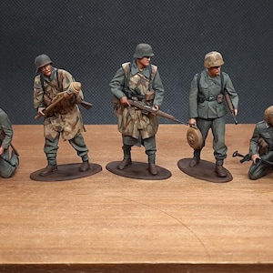May include: Five miniature military figures in various poses, dressed in olive green and camouflage uniforms, with helmets and weapons. The figures are standing on brown oval bases, set against a dark background.