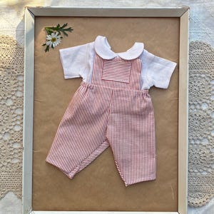 May include: A short-sleeved white shirt with a rounded collar layered under red and white vertically striped dungarees. The dungarees feature a rectangular chest pocket and the set is displayed on a flat surface with a small white flower.