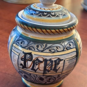 May include: A ceramic jar with a lid, decorated with blue, green, yellow, and brown floral patterns. The jar is labeled "Pepe" in black lettering.