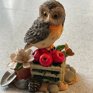 May include: A ceramic owl figurine perched on a wooden crate with red apples. The owl has brown and gray feathers and large, round eyes. The crate is surrounded by green leaves and a small flower pot with a white flower.