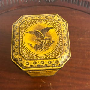May include: A yellow octagonal metal tin featuring a black graphic of an eagle in flight on the lid. The lid is decorated with intricate black scrollwork and circular patterns along the edges. The tin has a weathered finish with visible wear.