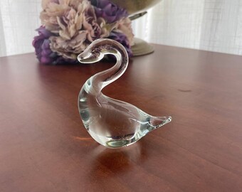 May include: A clear glass swan figurine with a long neck and a curved body. The swan is sitting on a wooden surface.