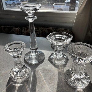 May include: Four clear glass candle holders of varying heights and designs. The tallest is a slender, stemmed candlestick. The others are shorter, with faceted bases and decorative tops. The candle holders are arranged on a gray surface.