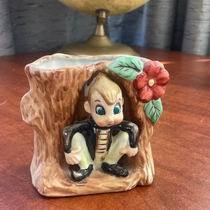 Puede incluir: Un macetero de cerámica con forma de tronco de árbol con una pequeña figura de gnomo sentada en su interior. El gnomo lleva una chaqueta marrón, pantalones blancos y zapatos negros. El macetero tiene una flor roja con hojas verdes en el lateral.
