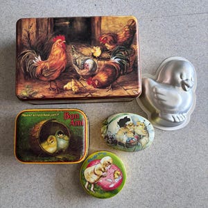 May include: A collection of vintage Easter-themed tins and a duck-shaped baking mold. The rectangular tin features a rooster and hens. Other tins have chick and egg designs. The duck mold is made of silver-colored metal.