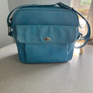 May include: A vintage, turquoise blue shoulder bag with a rectangular shape. The bag features a front flap with a metal clasp and a long adjustable strap. The bag is sitting on a light gray surface.