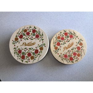 May include: Two round vintage cosmetic compacts with floral designs. The lids are off-white with a ring of red, orange, and green flowers. The word "Deluxe" is written in cursive across the center of each compact.