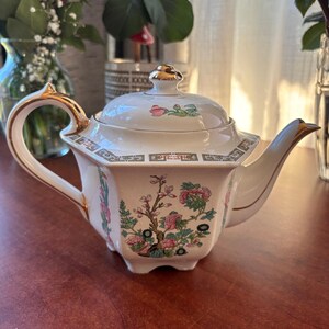 May include: White porcelain teapot with a floral design in pink, green, and gold accents. The teapot has a gold handle, spout, and lid finial. The floral pattern includes branches and flowers. The teapot is on a wooden surface.