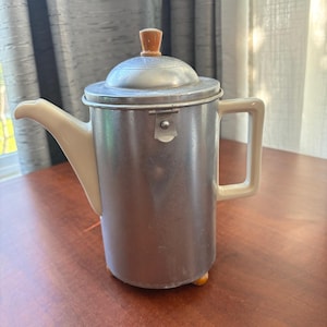 May include: A vintage metal coffee pot with a lid and a handle. The pot has a silver body, a cream-colored spout and handle, and a wooden knob on the lid. It sits on a wooden surface.