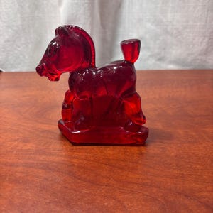May include: A translucent red glass rocking horse figurine. The horse is in a seated position with a rectangular base. The figurine is on a wooden surface with a white curtain in the background.