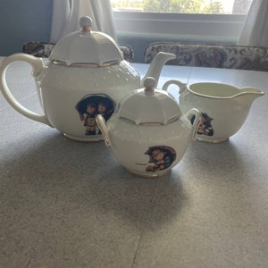 May include: A white porcelain tea set with a gold trim. The teapot, creamer, and sugar bowl feature a charming illustration of two children under an umbrella.