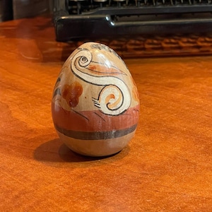 May include: A hand-painted ceramic egg with a brown, orange, and white design. The egg features a stylized bird design with a long neck and a curved beak.