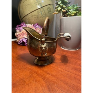 May include: A small brass coal scuttle with a lion head design and a white porcelain handle. The scuttle is on a wooden surface.