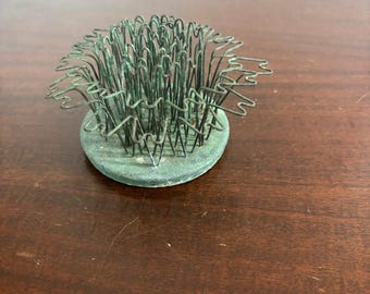 May include: A circular flower frog featuring a weathered green metal base and a series of upright, interconnected wire loops forming a cage-like structure. The metal components exhibit a patina with visible oxidation and signs of age and wear.