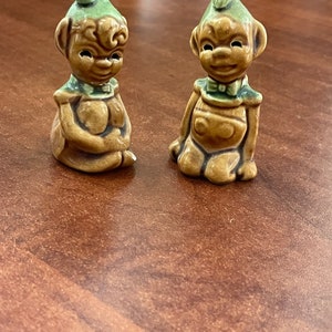 May include: Two ceramic figurines of elves with green hats and brown bodies. The elves are sitting with their legs crossed and have a whimsical expression on their faces.