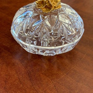 May include: A clear, cut-glass trinket dish with a gold-colored floral design on the lid. The dish has a round shape and a detailed, textured pattern. The lid's floral accent adds a touch of elegance to this decorative item.