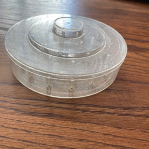 May include: A clear plastic round container with a lid. The container has a glittery finish and a circular design on the lid.