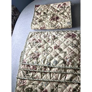 May include: A set of rectangular quilted placemats featuring a cream-colored background with a floral pattern in shades of burgundy, muted green, and light blue. The fabric has a diamond-stitched texture and is finished with a thin striped border.