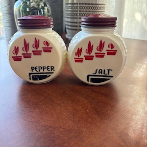 May include: Two vintage salt and pepper shakers. The white, round shakers have a textured surface and a maroon lid. Each shaker has a red design of potted plants. The word "PEPPER" is printed on one shaker, and "SALT" on the other.