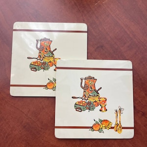 May include: Two white ceramic trivets with brown trim. Each trivet features a colorful illustration of a coffee pot, fruit, and kitchen utensils.