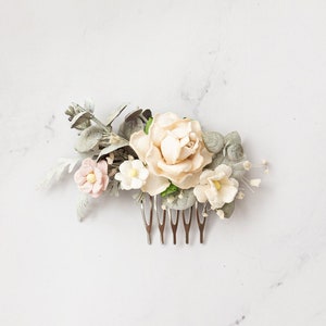 Puede incluir: Un peine para el cabello floral con una gran rosa color melocotón, pequeñas flores rosas y blancas y hojas de color verde grisáceo. Las flores están unidas a un peine plateado, ideal para bodas u ocasiones especiales.