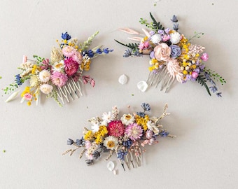 May include: Three floral hair combs with dried flowers in shades of pink, yellow, blue, and white. The combs are arranged on a white surface.