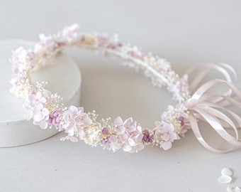 May include: A delicate floral crown featuring light pink and purple flowers, accented with small white blossoms. The crown is tied with a soft pink ribbon. Perfect for weddings or special occasions.