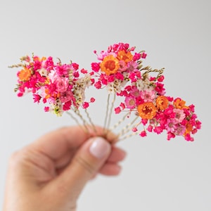 Peut inclure: Plusieurs épingles à cheveux florales sont tenues dans une main. Chaque épingle présente un bouquet de petites fleurs vibrantes dans les tons de rose, d'orange et de blanc. Les fleurs sont fixées à des épingles à cheveux dorées, créant un accessoire délicat et coloré.