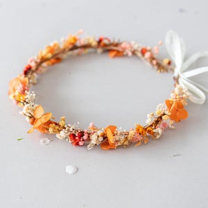 Könnte beinhalten: Eine Trockenblumenkrone mit orangefarbenen und weißen Blüten. Die Krone besteht aus einem dünnen Drahtgestell und hat ein weißes Band.