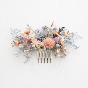 May include: A floral hair comb featuring a variety of dried flowers in shades of peach, orange, lavender, and blue. The flowers are arranged on a silver comb with six teeth. The comb is a decorative hair accessory.