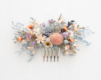 May include: A floral hair comb featuring a variety of dried flowers in shades of peach, orange, lavender, and blue. The flowers are arranged on a silver comb with six teeth. The comb is a decorative hair accessory.