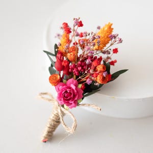 Könnte beinhalten: Eine lebendige florale Ansteckblume mit einer rosa Rose, orangefarbenen und roten Blüten und grünen Blättern. Die Blumen sind mit Bindfaden gebunden, wodurch ein rustikales und elegantes Accessoire entsteht. Perfekt für Hochzeiten oder besondere Anlässe.