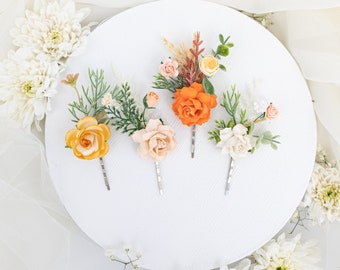 Puede incluir: Cuatro pinzas para el pelo florales con flores de diferentes colores. Las pinzas presentan una rosa melocotón, una rosa naranja, una rosa amarilla y una rosa blanca con vegetación.
