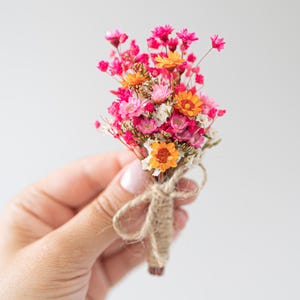 Op de afbeelding: Een kleine boeket van gedroogde bloemen in roze, oranje en wit, samengebonden met touw. De bloemen zijn compact gerangschikt en tonen een verscheidenheid aan texturen en kleuren. De achtergrond is een zachte, neutrale tint.