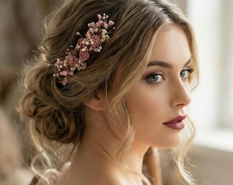 May include: A woman with blonde hair styled in an updo, adorned with a floral hairpiece. The hairpiece features small pink and white flowers. She is wearing a white dress and sitting in a wooden chair.