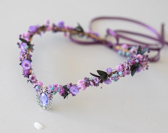 May include: A floral crown featuring a delicate arrangement of purple and pink dried flowers, accented with small black leaves. A silver pendant with a purple gemstone hangs at the front. The crown has purple ribbons for tying.
