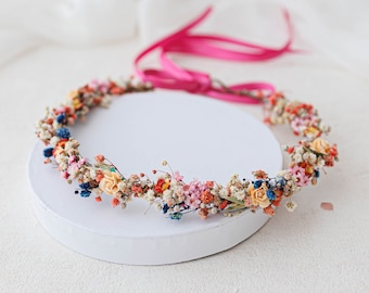 May include: A floral crown made of dried flowers in shades of pink, orange, and blue. The crown is on a white circular stand.