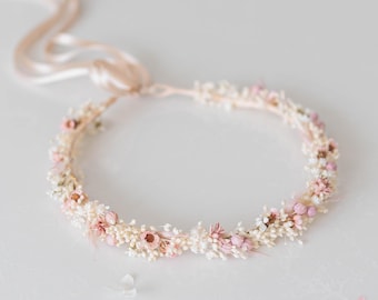 Könnte beinhalten: Eine zarte Blumenkrone aus getrockneten weißen und rosa Blüten. Die Krone ist mit einem hellrosa Band gebunden.