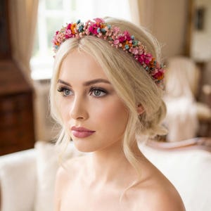 Könnte beinhalten: Eine Frau mit blonden Haaren trägt einen Blumenkranz mit rosa, orangefarbenen und blauen Blüten. Sie trägt ein weißes trägerloses Kleid. Im Hintergrund sind ein weißes Sofa und ein Fenster mit transparenten Vorhängen zu sehen.
