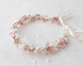 May include: A delicate floral crown made with dried flowers in shades of white and blush pink. The crown is made with a variety of small flowers and greenery, and is tied with a white satin ribbon.