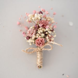 Könnte beinhalten: Ein kleiner, handgefertigter Anstecker mit getrockneten Blumen in Rosa, Creme und Burgunder. Die Stiele sind mit Bindfaden umwickelt und mit einer zarten Schleife gebunden. In der Mitte befindet sich eine einzelne, zarte rosa Rose.
