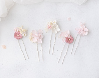 May include: Five floral hair pins in shades of pink and white are arranged on a white surface. Each pin features delicate fabric flowers and small, pearl-like accents. The pins are ideal for weddings or special occasions.