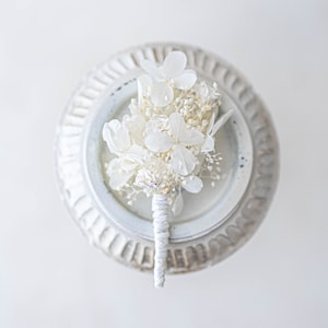May include: A white dried flower bouquet with a white ribbon, displayed on a white pedestal.
