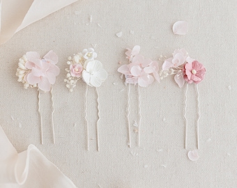 May include: Four delicate floral hair pins with pink and white flowers. The pins are arranged in a row on a light beige fabric background.