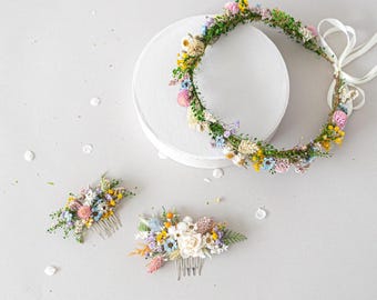 May include: A set of three floral hair accessories, including a flower crown and two hair combs. The crown is made of dried flowers in shades of pink, yellow, blue, and white, and is tied with a white ribbon. The hair combs are also decorated with dried flowers in similar colors.