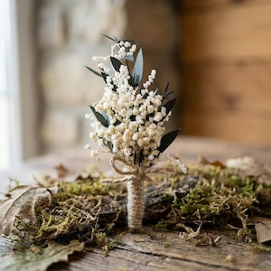Op de afbeelding: Een kleine, handgemaakte corsage met delicate witte bloemen en donkergroene bladeren. De stelen zijn omwikkeld met touw en vastgebonden met een kleine strik. De corsage ligt op een bedje van mos en bladeren, wat een rustiek of natuurlijk thema suggereert.