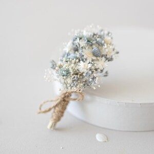 May include: A small, dried flower bouquet featuring white, light blue, and green blossoms. The stems are wrapped with twine and tied with a small bow. The bouquet is a delicate, natural accent for weddings or other special events.